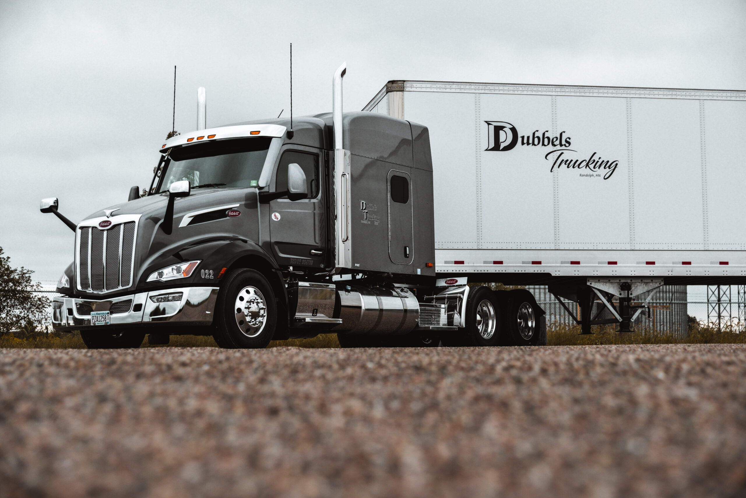 A gray semi-truck with a white trailer marked Dubbels Trucking is parked on a gravel surface under a cloudy sky.
